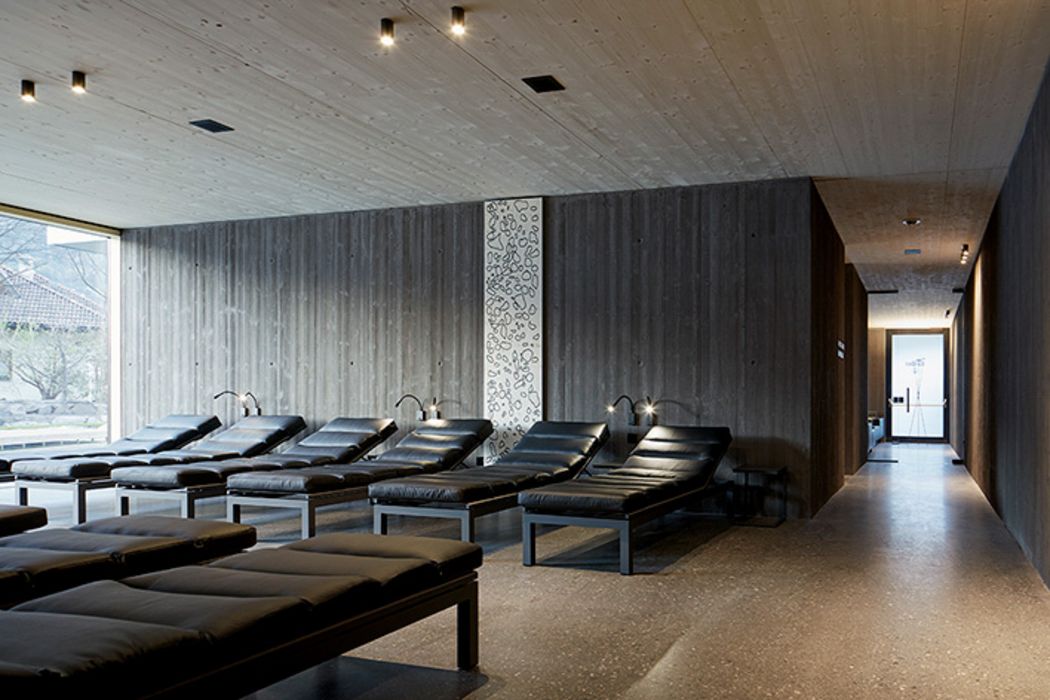 Ein moderner, minimalistischer Ruheraum in einem Spa mit einer Reihe schwarzer Lederliegen vor einer grauen Sichtbetonwand und einem dekorativen, gemusterten Paneel.