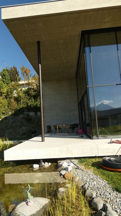Eine überdachte moderne Betonveranda mit einem Gartentisch und Stühlen vor einer Glasfassade, angrenzend an einen kleinen Teich.