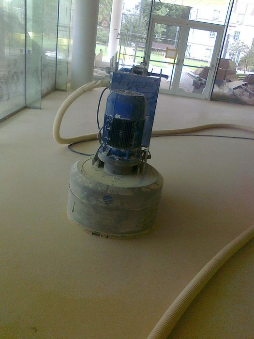 Eine Boden-Schleifmaschine mit blauem Motor steht auf einem staubigen, neu geschliffenen Betonboden in einem Gebäude mit Glasfront.