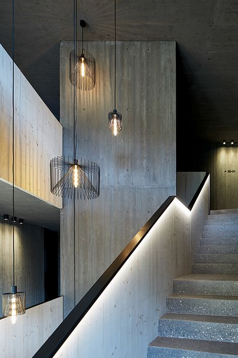 Innenansicht eines Treppenhauses mit Betonwänden, Holzverkleidung und modernem Treppengeländer mit integrierter LED-Beleuchtung, von der Decke hängen mehrere schwarze Drahtnetz-Pendelleuchten.