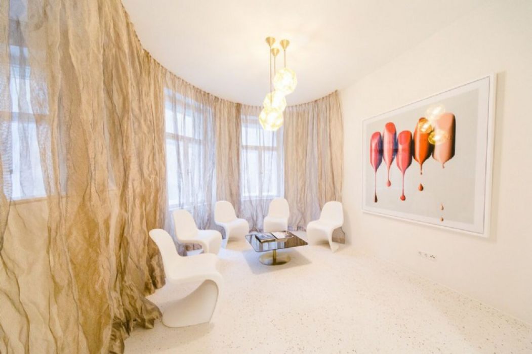 Ein heller Wartebereich mit vier weißen geschwungenen Stühlen, die um einen kleinen Couchtisch angeordnet sind, vor einer Fensterwand mit halbtransparenten beigen Vorhängen und einem großen Kunstbild mit Lippenstiften an der Wand.