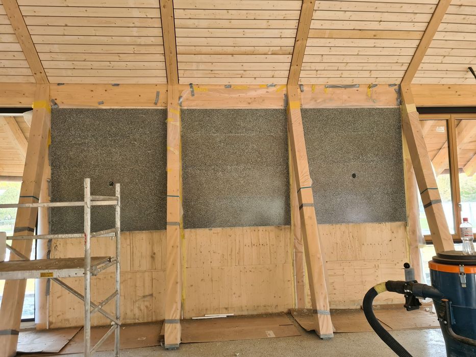 Innenansicht einer Holzbaukonstruktion, bei der graue Steinplatten zwischen hölzerne Stützpfeiler eingebaut wurden, mit einem Gerüst links und einem Baustellenstaubsauger rechts.