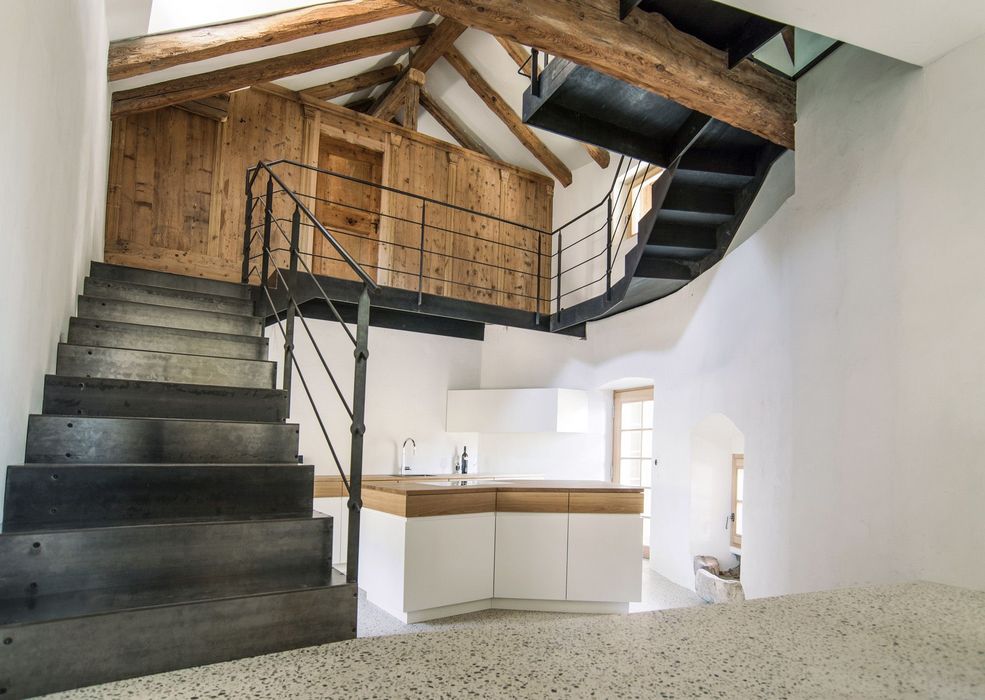 Innenraum eines modernen Hauses im Dachgeschoss mit einer schwarzen Metalltreppe, einer Kochinsel mit Holzelementen und freiliegender Holzbalkendecke.