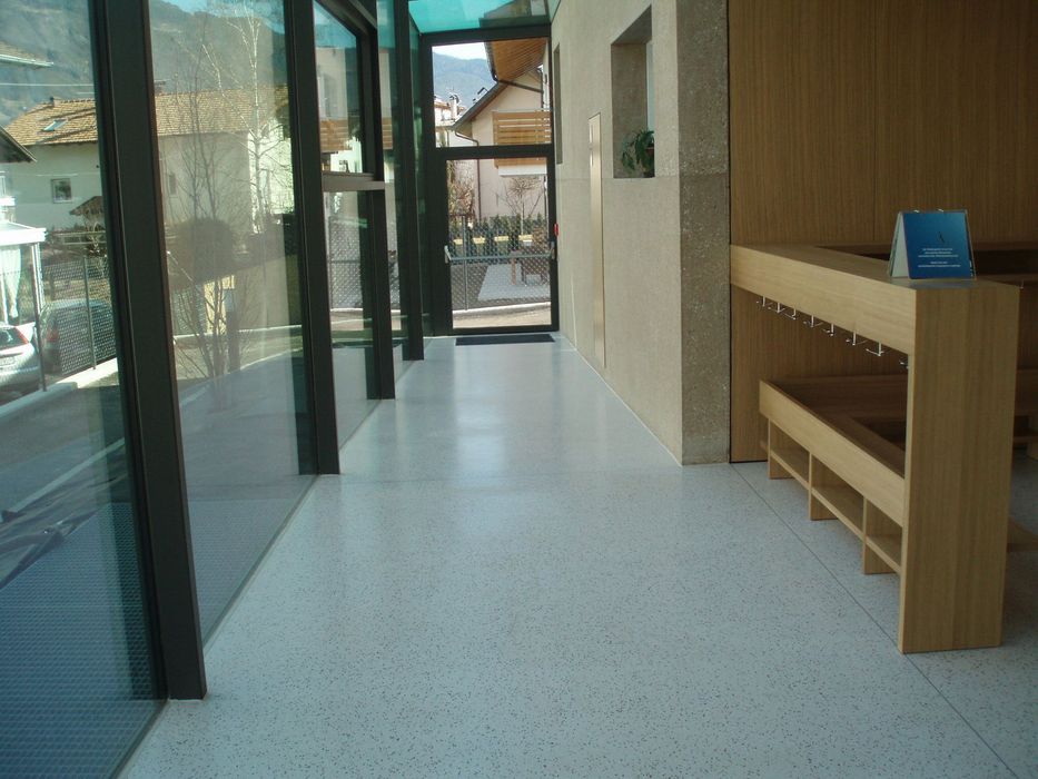 Ein heller, moderner Flur mit einer großen Glasfront links, einem hellen gesprenkelten Boden und einer integrierten Garderobe aus Holz rechts, der Blick führt auf eine Tür im Hintergrund.