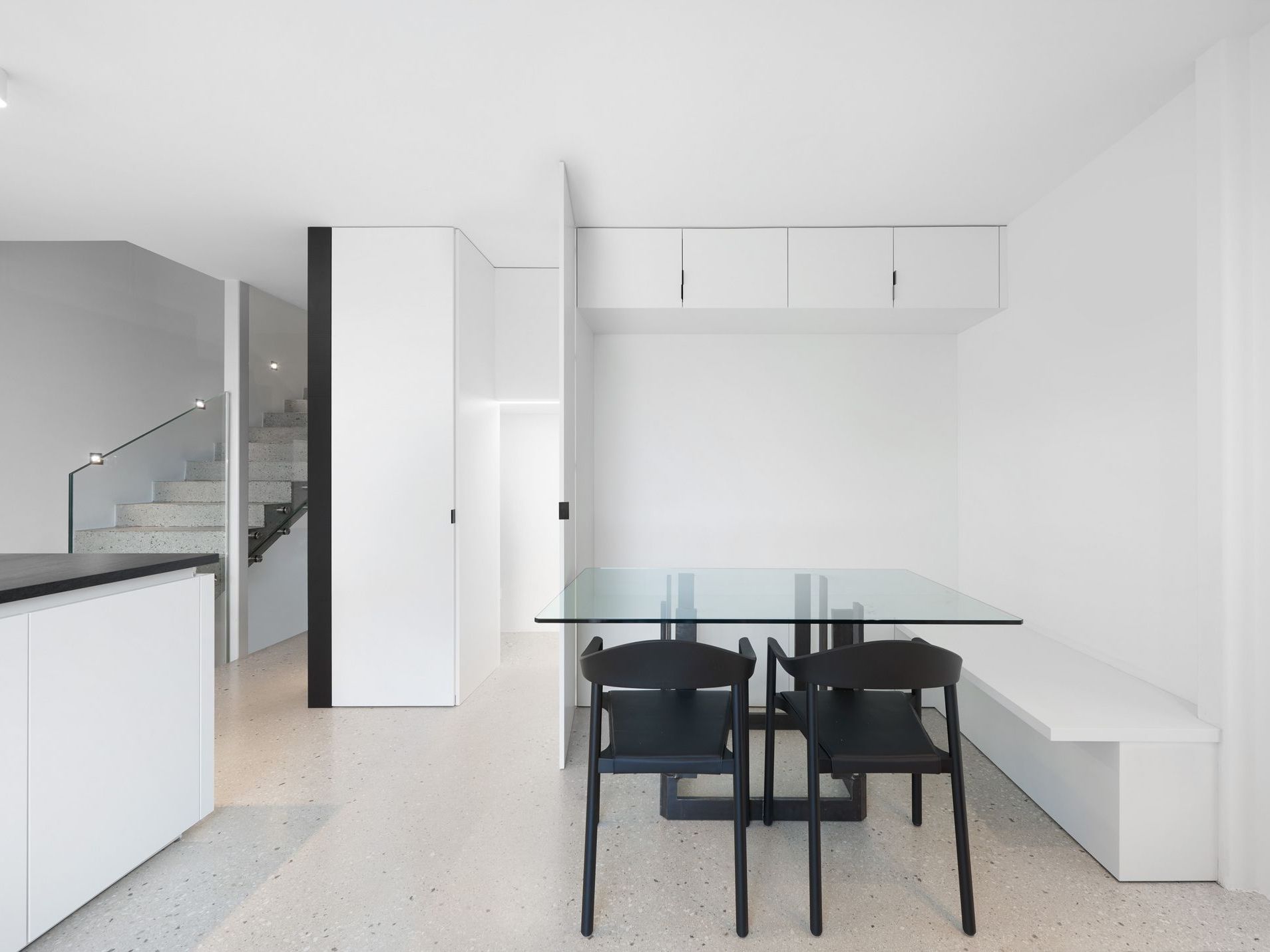 Ein minimalistischer Essbereich mit einem Esstisch aus Glas und zwei schwarzen Stühlen, flankiert von weißen Einbauschränken und einer Treppe im Hintergrund.