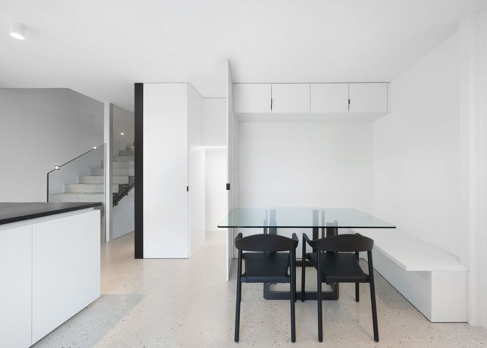 Ein minimalistischer Essbereich mit einem Esstisch aus Glas und zwei schwarzen Stühlen, flankiert von weißen Einbauschränken und einer Treppe im Hintergrund.