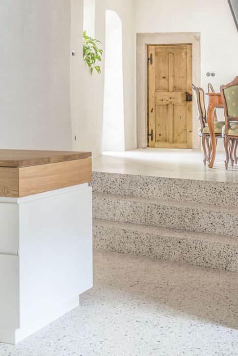 Ein heller Innenraum mit einem weißen Küchenblock im Vordergrund, der über eine Treppe aus Terrazzo-Fliesen zu einem tiefer gelegenen Bereich mit einem antiken Holztisch und Stühlen führt.