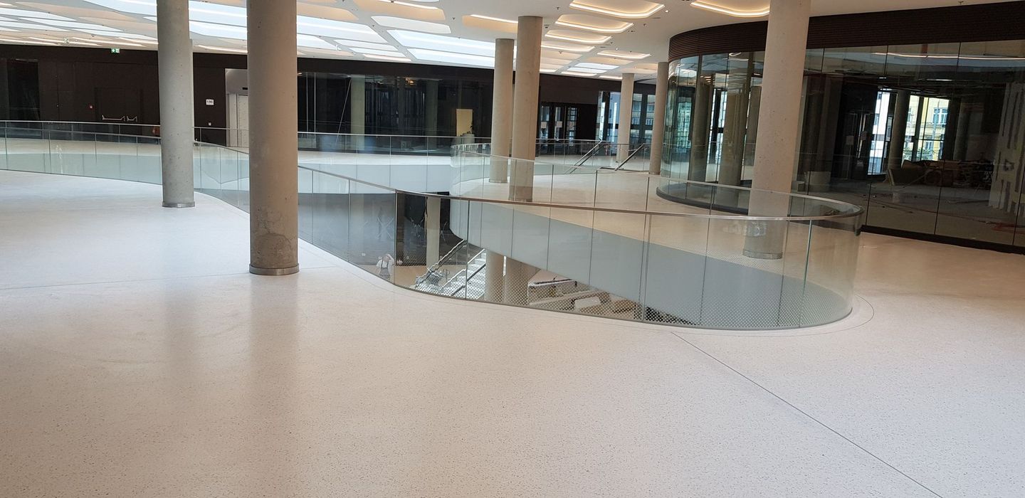 Ein minimalistisches, helles Atrium mit geschwungenen Glasgeländern, Betonstützen und glatten Bodenbelägen in einem modernen Gebäude.