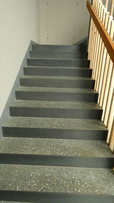 Eine gerade Treppe aus grauem Terrazzo-Stein führt nach oben, mit einem schlichten Holzgeländer auf der rechten Seite.