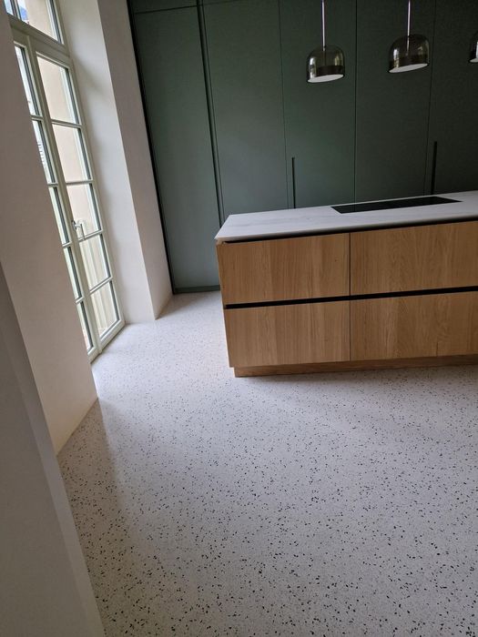 Eine moderne Küche mit einer Kücheninsel aus Holz, einer weißen Arbeitsplatte und dunkelgrünen Schrankfronten auf einem hellen Terrazzoboden.