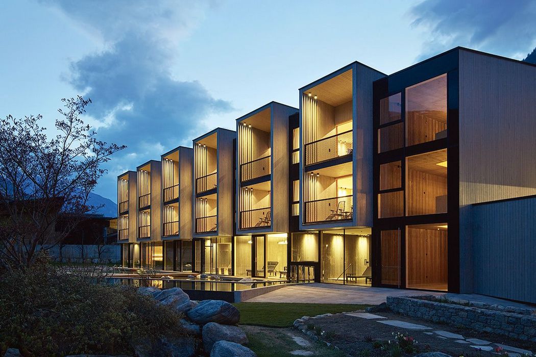 Ein modernes Hotelgebäude aus Holz und Glas in der Abenddämmerung mit beleuchteten Balkonen und einem kleinen Poolbereich im Vordergrund.