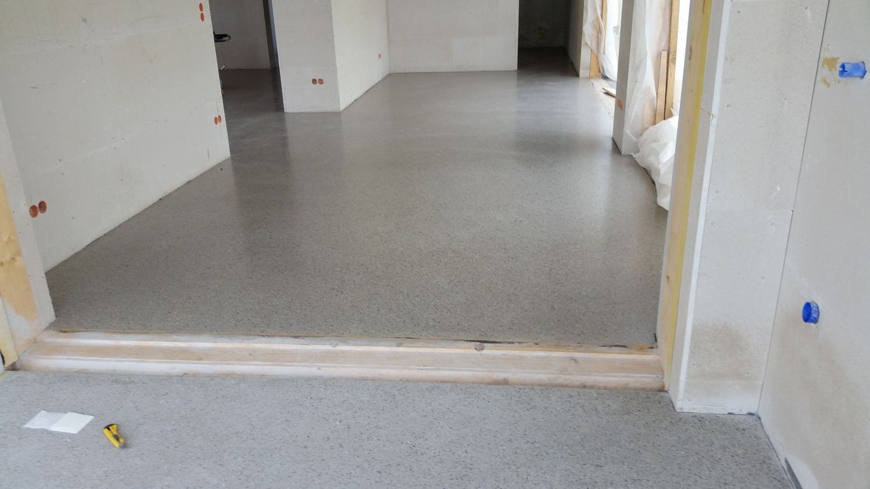 Ein Rohbau-Innenraum mit frisch verlegtem, glattem Estrichboden zwischen weiß verputzten Wänden und einem hölzernen Türrahmen.