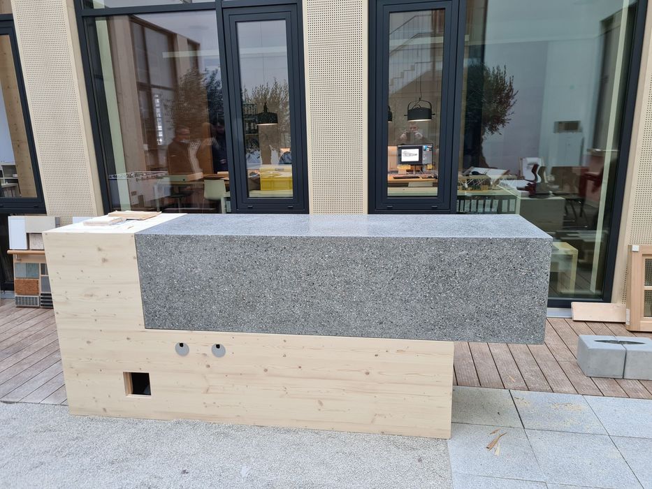 Ein länglicher Tresen mit einem Unterbau aus hellem Holz und einem darüber schwebenden rechteckigen Block aus grauem Terrazzo vor einer Glasfront.