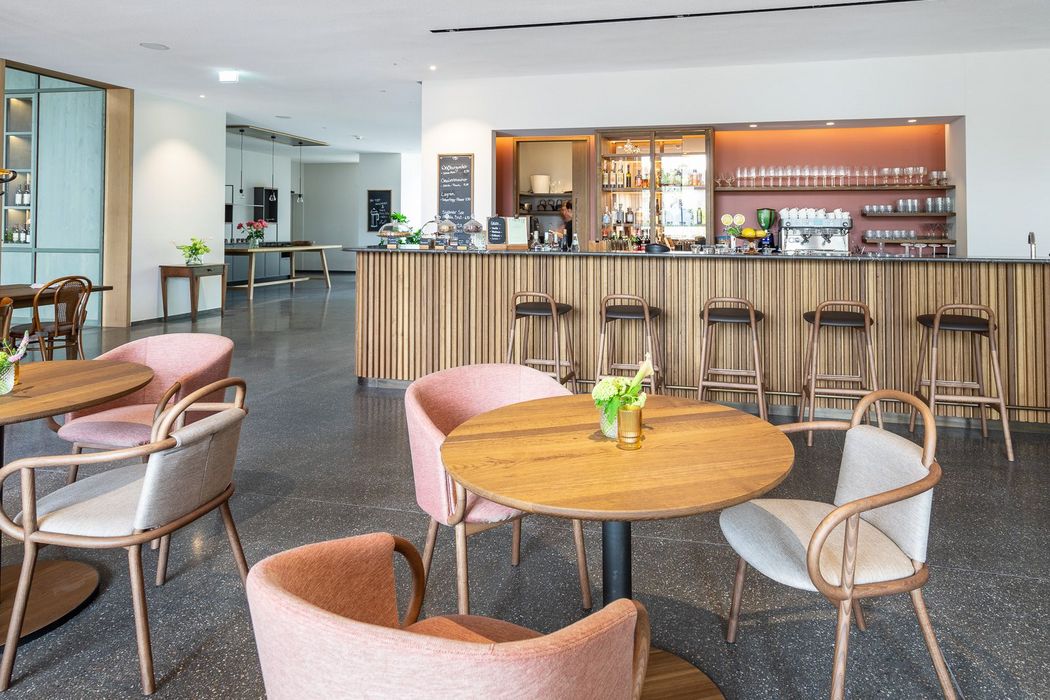 Innenansicht eines modernen Cafés mit runden Holztischen, gepolsterten Stühlen in Rosa und Grau und einem langen Tresen mit Barhockern vor einer roten Wand.