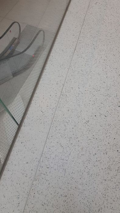 Ein gesprenkelter heller Terrazzo-Boden in Innenräumen, neben dem eine gläserne Rolltreppenkonstruktion zu sehen ist.