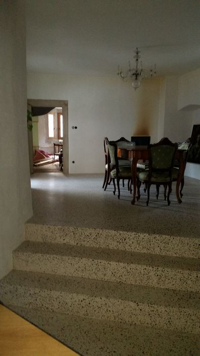 Ein Blick durch eine architektonische Öffnung auf ein Esszimmer mit einem hölzernen Tisch und grünen Polsterstühlen, ergänzt durch einen Kronleuchter an der hohen Decke und einen Terrazzo-Treppenaufgang im Vordergrund.