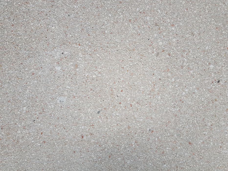 Nahaufnahme einer grauen Betonoberfläche mit feiner, gesprenkelter Körnigkeit und kleinen rötlichen Gesteinseinschlüssen.