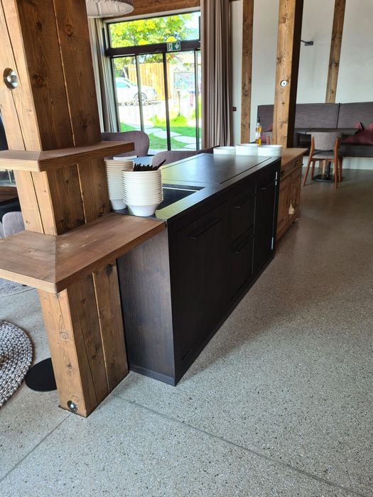 Ein dunkler Tresen in einem Gastraum aus Holz mit gestapelten weißen Schalen, im Hintergrund sind Tische, Stühle und eine große Glasfront mit Blick nach draußen zu sehen.