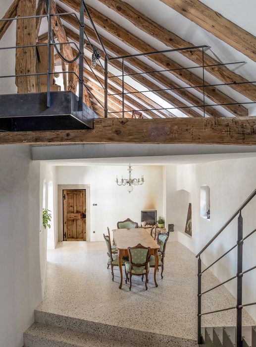 Ein Blick von oben auf einen hellen Essbereich mit rustikalen Holzbalken, einem antiken Holztisch mit grünen Stühlen und einer Steintreppe.