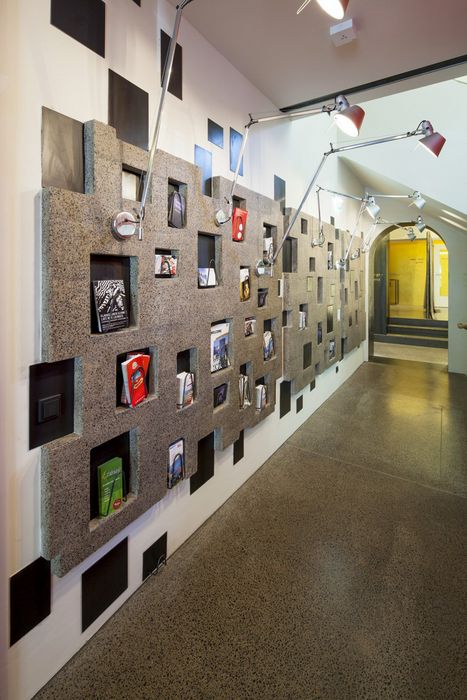 Ein langer Flur mit einem Terrazzo-Boden und einer Wandgestaltung aus strukturierten Steinpaneelen, in die Aussparungen eingelassen sind, in denen Broschüren und Informationsmaterial stehen.