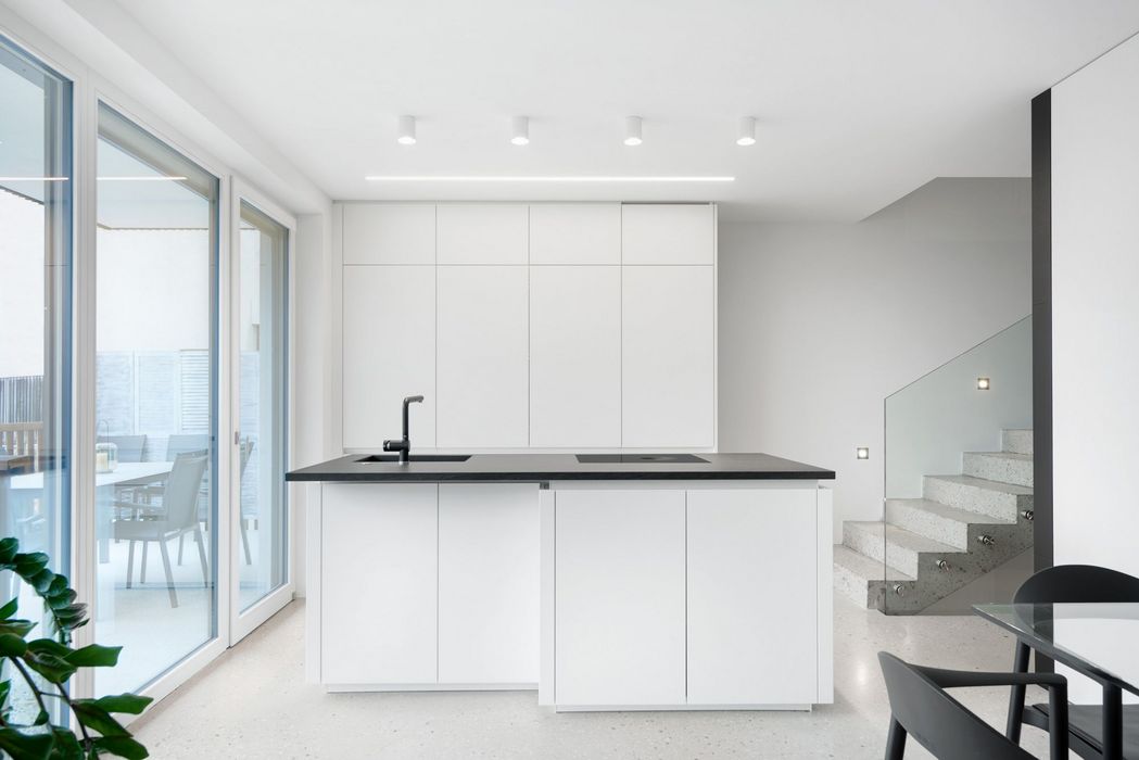 Eine moderne, minimalistische weiße Küche mit einer schwarzen Kochinsel und schwarzer Armatur in einem hellen, offenen Wohnbereich, der an eine Terrasse grenzt und neben einer Treppe mit Glasgeländer liegt.