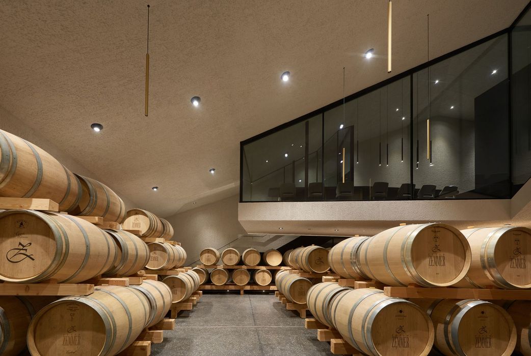 Ein modern gestalteter Weinkeller mit zahlreichen aufgestapelten Holzfässern der Marke Peter Zemmer auf einem Steinboden, daneben eine durch Glas abgetrennte Ebene mit einer Treppe und Beleuchtung.