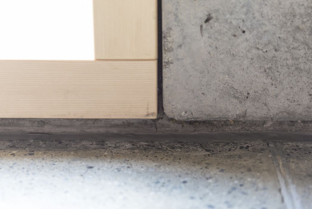 Nahaufnahme eines Fensterrahmens aus hellem Holz, der direkt auf einer rauen Betonoberfläche mit einer Fuge aus Silikon oder Dichtstoff montiert ist.
