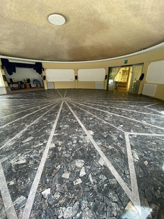 Ein runder Festsaal mit einem dunkelgrauen Steinboden, der eine sternförmige Verzierung aufweist, und einer kleinen Bühne mit blauem Vorhang.