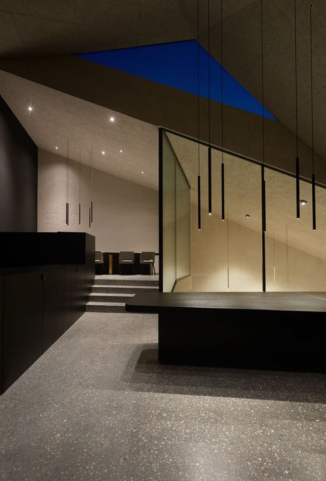 Ein minimalistisch gestalteter Innenraum mit grauen Terrazzoböden, einer schwarzen Insel, Stufen zu einem Essbereich und schmalen, hängenden Beleuchtungselementen unter einem schrägen Dach mit sichtbarem blauem Abendhimmel.