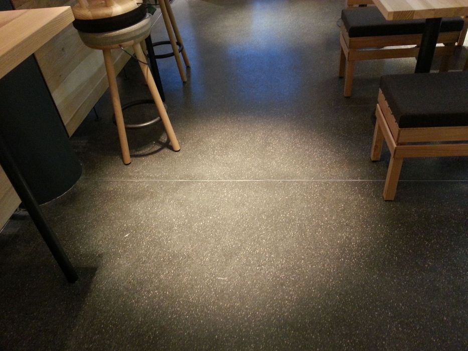 Innenraum eines Cafes mit Holzstühlen und Barhockern auf einem dunkelgrau gesprenkelten Boden.