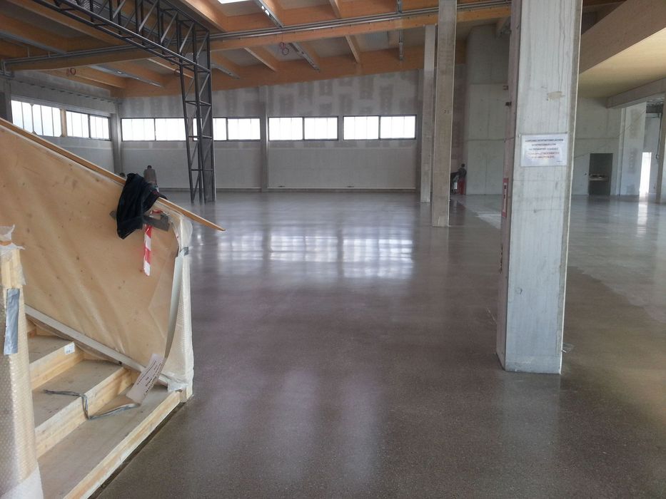 Innenansicht einer großen, leeren Halle während der Bauphase mit glattem Betonboden, Holzbalkendecke und einer Treppenkonstruktion im Vordergrund.