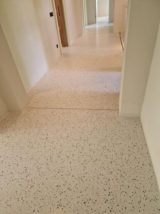 Ein heller Flur mit einem modernen weißen Terrazzo-Boden mit dunklen Sprenkeln und weißen Wänden, der in einen anderen Raum führt.