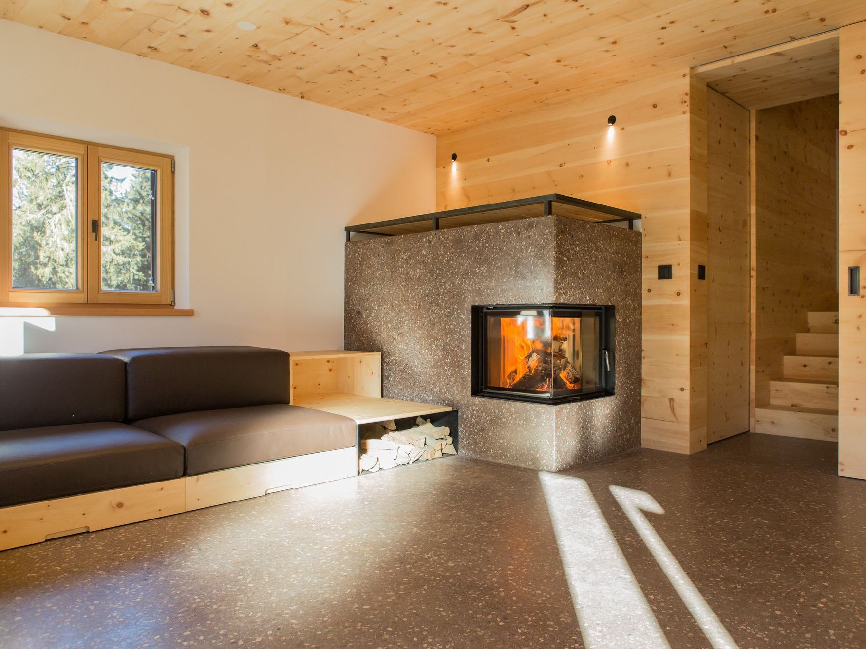 Ein minimalistisch eingerichteter Wohnraum mit einem modernen Eckkamin, einem dunkelgrauen Sofa mit integriertem Holzregal und holzverkleideten Wänden und Decken.