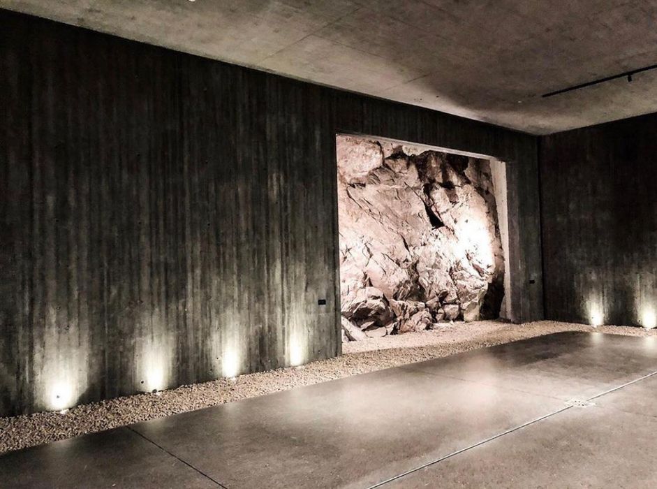 Ein moderner Raum mit Sichtbetonwänden und Bodenbeleuchtung, der an einer der Wände durch ein Fenster eine unberührte, beleuchtete Felsformation freigibt.
