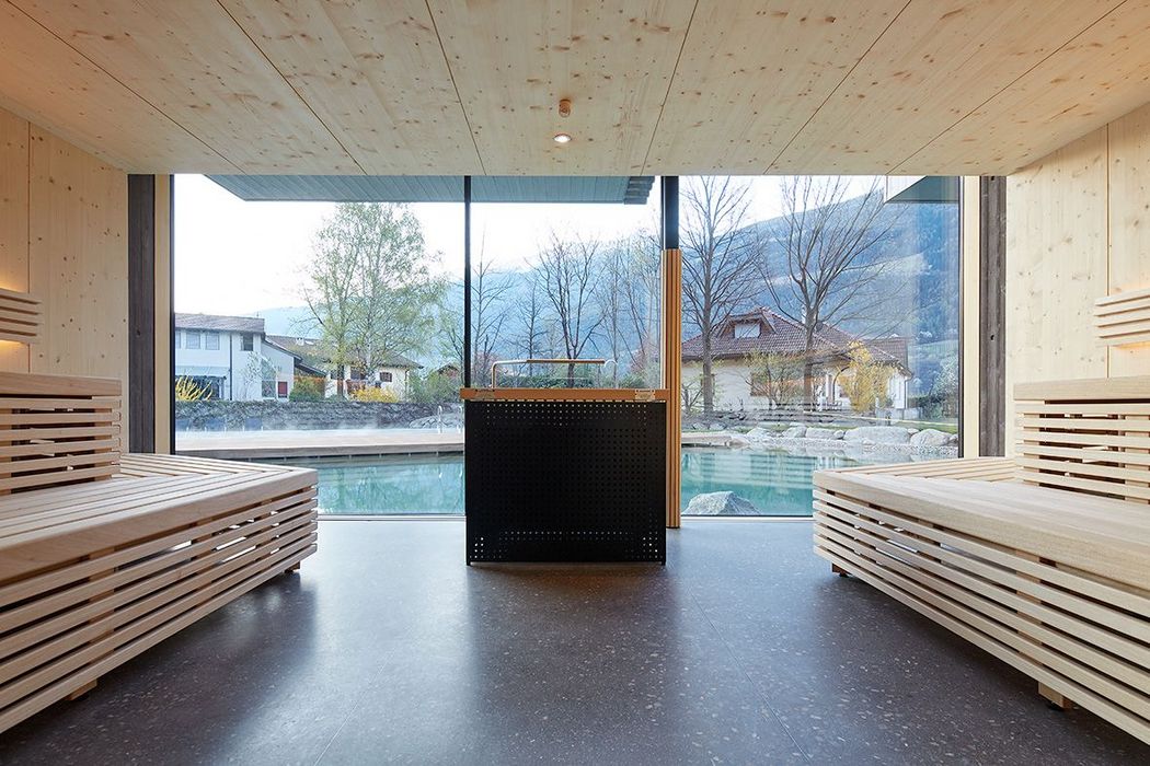 Innenansicht einer modernen holzverkleideten Sauna mit zwei gegenüberliegenden Holzbänken und einem großen Panoramafenster, das den Blick auf einen Außenpool und eine bewaldete Hügellandschaft freigibt.
