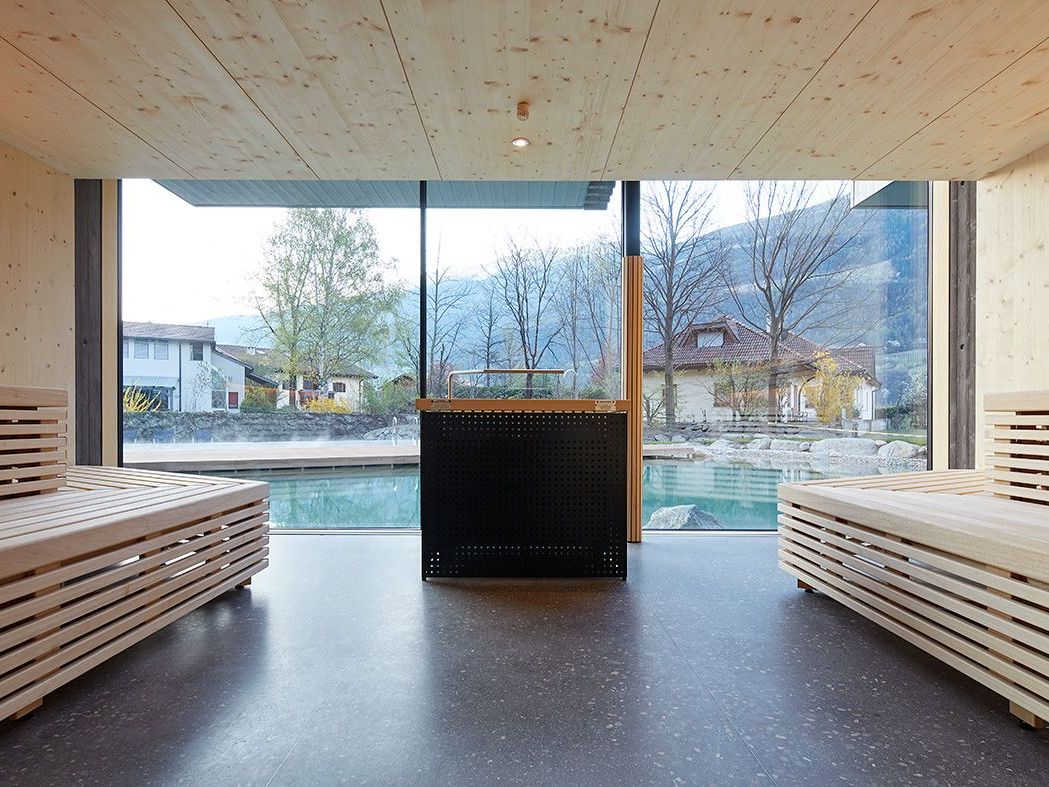 Innenansicht einer modernen holzverkleideten Sauna mit zwei gegenüberliegenden Holzbänken und einem großen Panoramafenster, das den Blick auf einen Außenpool und eine bewaldete Hügellandschaft freigibt.