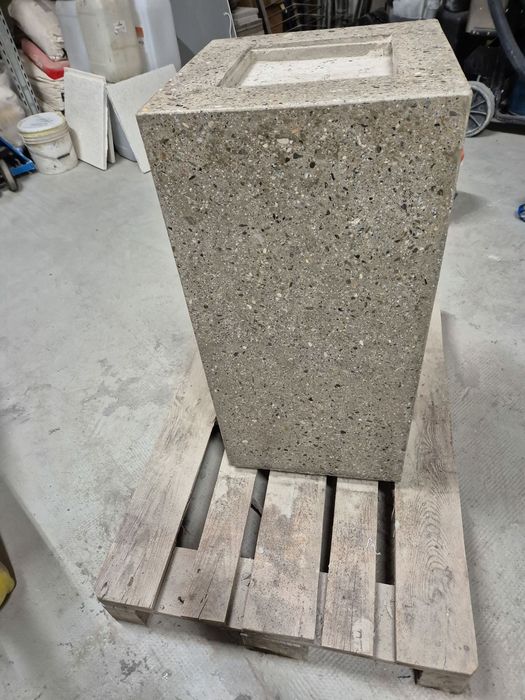 Ein rechteckiger Betonsockel mit gesprenkelter Oberfläche steht auf einer hölzernen Palette in einer Lagerhalle.