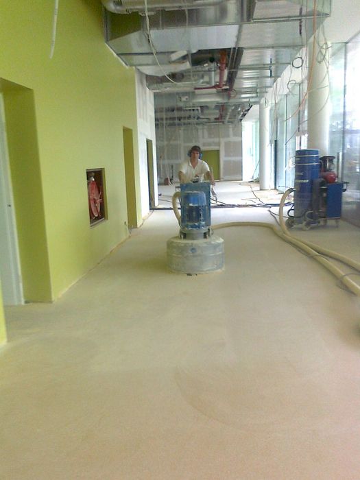 Ein Arbeiter bedient eine Bodenschleifmaschine in einem im Bau befindlichen Flur mit hellgrüner Wand und offenliegenden Deckeninstallationen.