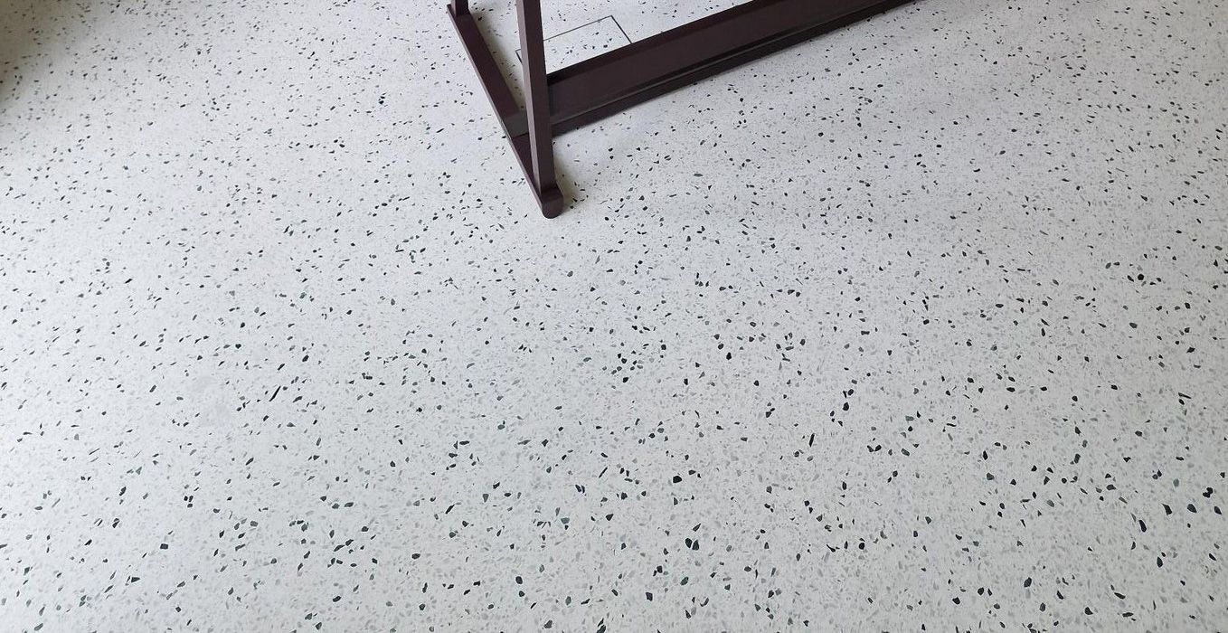 Ein heller Terrazzoboden mit kleinen schwarzen und grauen Sprenkeln, im oberen Bereich ist ein Teil eines dunklen Tischbeins zu sehen.