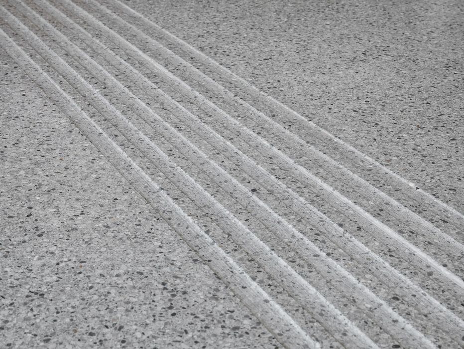 Ein grauer Steinboden mit einer diagonal verlaufenden Reihe von erhabenen Rillen für die taktile Orientierung.