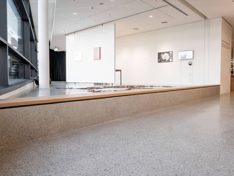 Ein minimalistischer Innenraum einer Kunstgalerie mit einer niedrigen Betonplattform, auf der Holzzweige liegen, sowie weißen Stellwänden mit verschiedenen gerahmten Bildern.