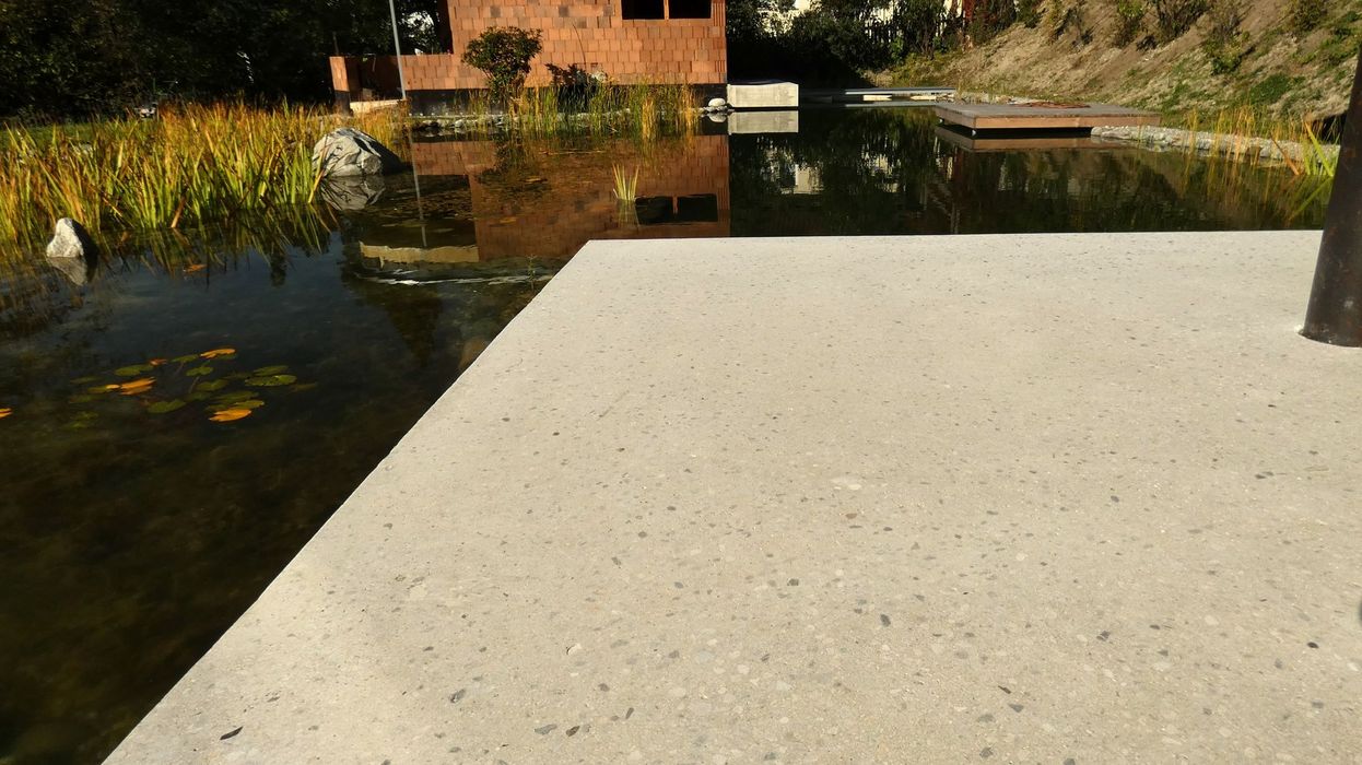 Eine helle Betonplattform ragt in einen naturnahen Gartenpool, in dessen ruhigem Wasser sich das Haus im Hintergrund spiegelt.