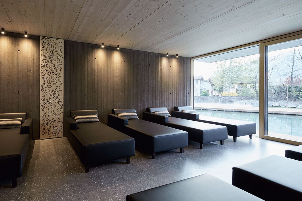 Ein moderner Ruheraum mit mehreren schwarzen Liegen vor einer texturierten Holzwand, neben einer großen Fensterfront mit Blick auf einen Außenpool.