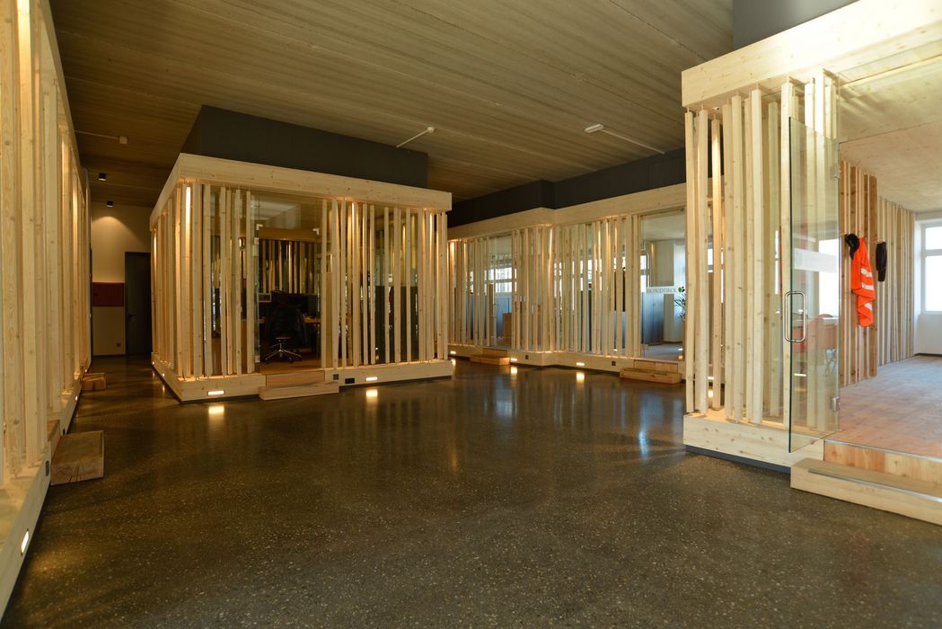 Ein moderner Innenraum mit mehreren freistehenden, kastenförmigen Büroeinheiten aus Holzlatten und Glaselementen auf einem dunklen Boden.