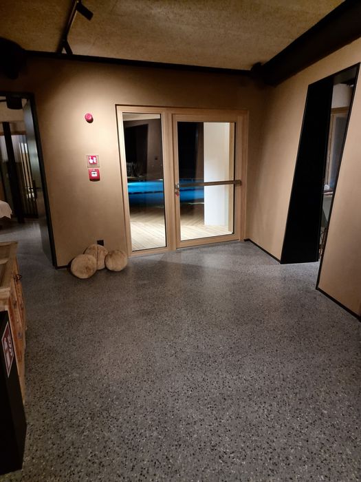 Ein Innenraum mit grauem Terrazzoboden, einer gläsernen Terrassentür, die den Blick auf einen beleuchteten Pool bei Nacht freigibt, und dekorativen runden Holzelementen in der Ecke.