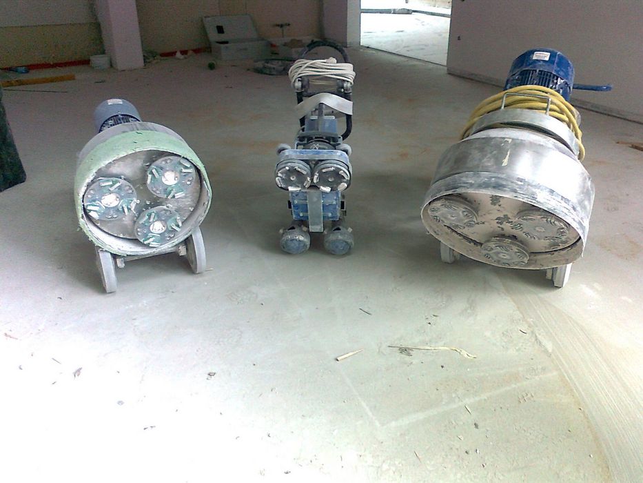 Drei Industriemaschinen zum Schleifen von Betonböden stehen auf einem staubigen Baustellenboden.