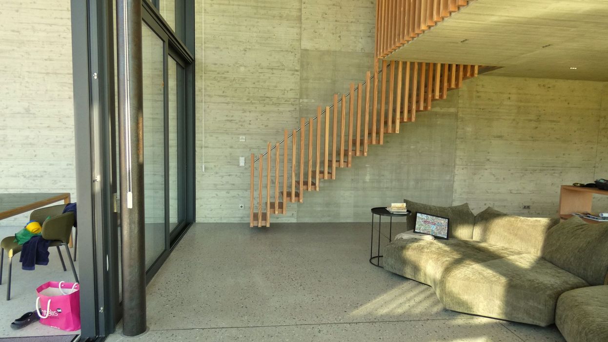 Ein minimalistischer Wohnraum mit Sichtbetonwänden, einer freischwebenden Holztreppe, einem Sofa und einer großen Glasfront.