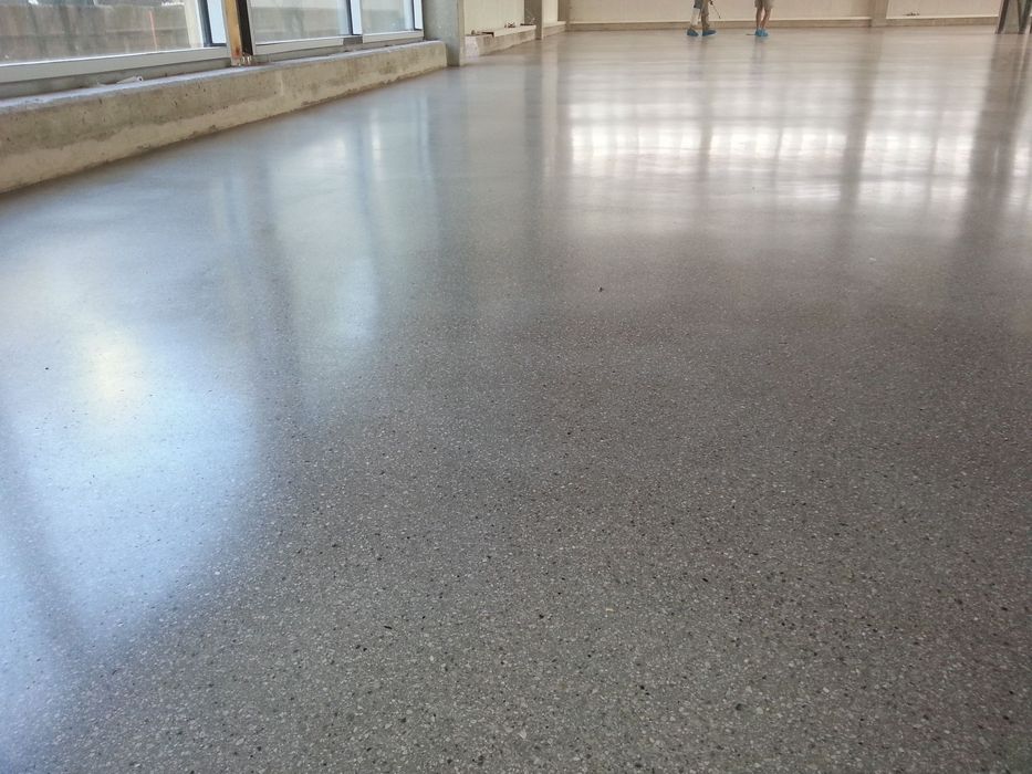 Ein frisch polierter, grauer Sichtbetonboden in einem hellen, leeren Raum mit großen Fenstern an der Seite und zwei Personen im Hintergrund.