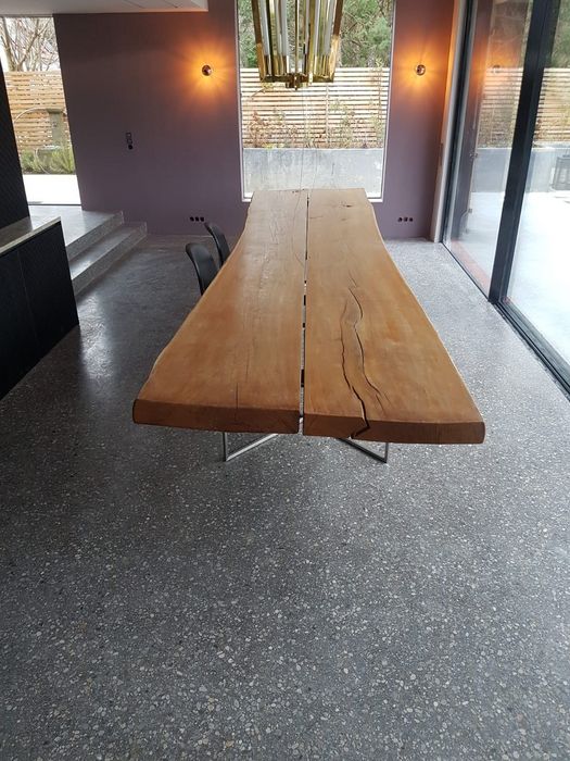 Ein langer Esstisch aus einer massiven, naturbelassenen Holzplatte steht in einem modernen Esszimmer mit Terrazzoboden, vor einer großen Fensterfront.