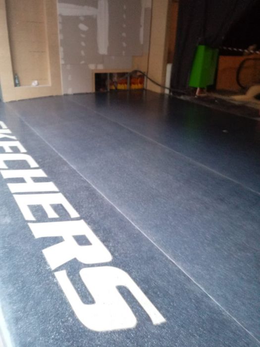 Ein dunkler Bodenbelag in einem Innenraum im Umbau, auf dem in großen weißen Buchstaben der Schriftzug SKECHERS zu sehen ist.